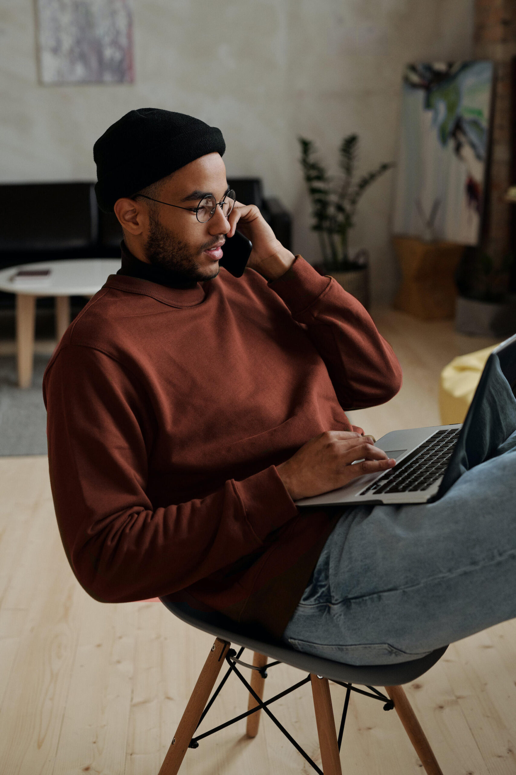 Jeune adulte garçon avec une tuque noir et un chandail marron parlant au téléphone assis avec son ordinateur portable sur ses cuisses.